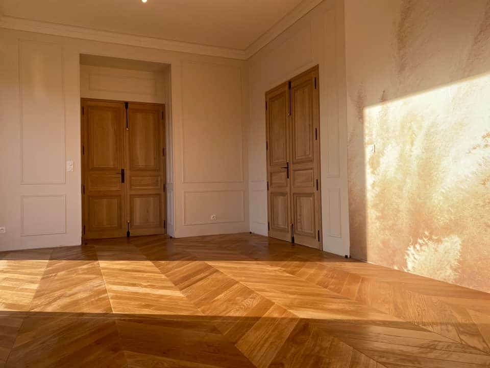 Intérieur élégant avec parquet à chevrons en bois clair, murs moulurés blancs et doubles portes en bois massif éclairées par un fort ensoleillement.