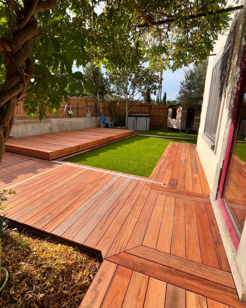 Terrasse bois rétractable et jardin moderne Terrasse en bois rouge moderne avec un panneau coulissant couvrant une piscine ou un espace de vie, juxtaposée à une pelouse synthétique verte.