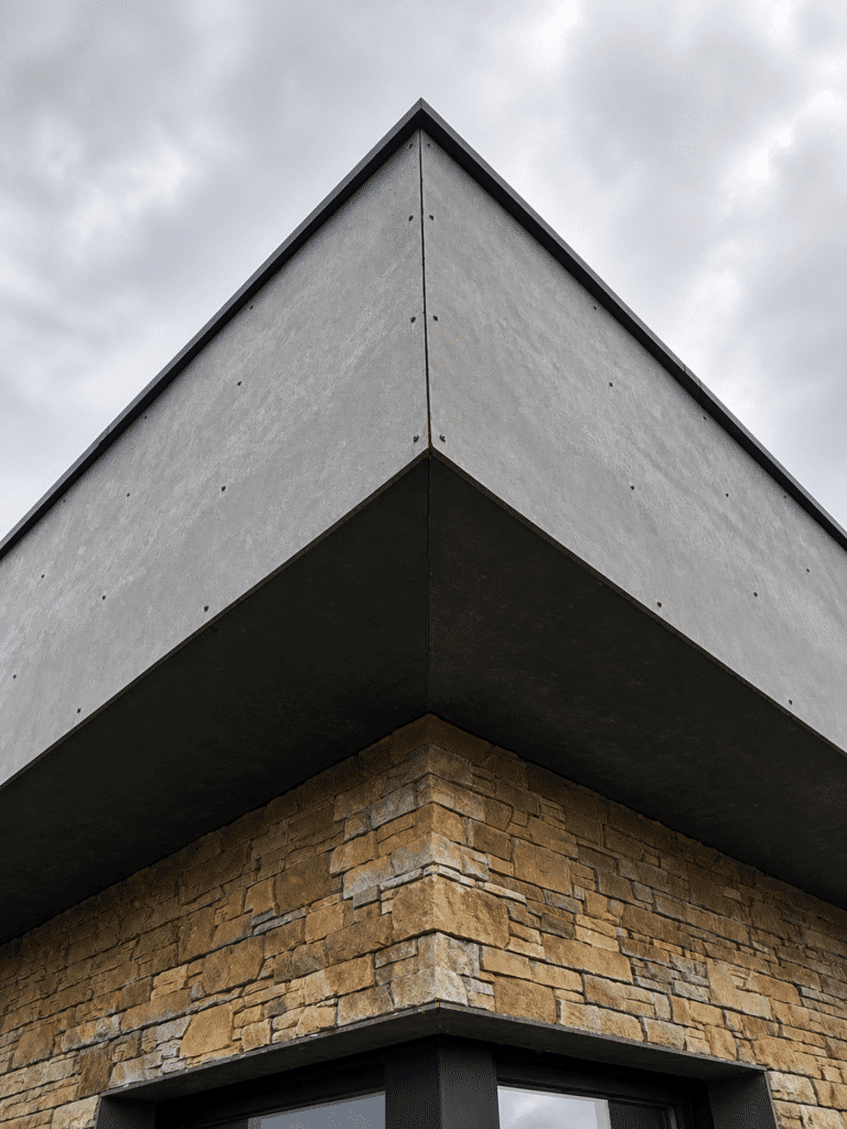 Architecture Moderne : Façade Pierre et Panneaux Gris Angle de bâtiment moderne alliant panneaux gris lisses et parement en pierre naturelle sous un ciel nuageux.