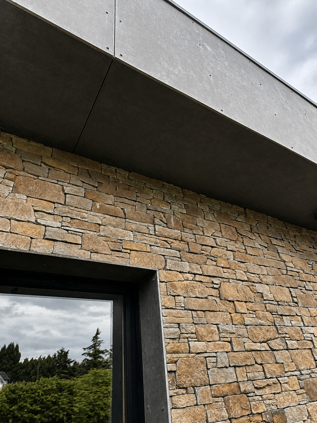 Parement en pierre naturelle et débord de toit gris anthracite au-dessus d'une fenêtre vitrée.