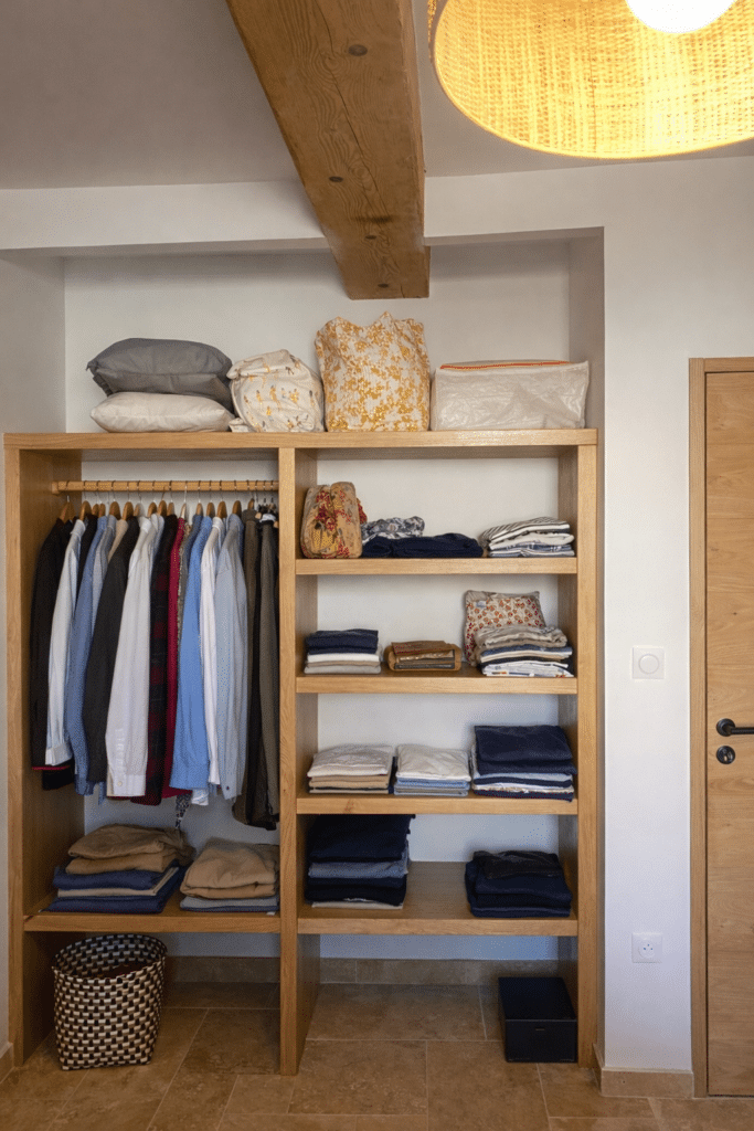 Dressing en bois avec chemises suspendues, vêtements pliés et panier tressé sous une poutre apparente.