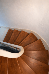 Escalier tournant en bois brun massif, rampe blanche et murs clairs en vue plongeante.