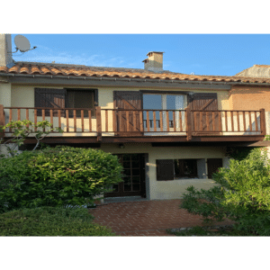 Maison provençale avec balcon en bois, volets bruns et terrasse en briques entourée de buissons verts.